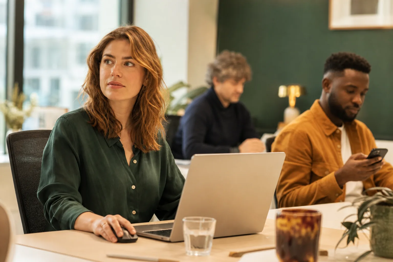 Kantoortuin met medewerkers achter laptops, op de voorgrond een vrouw die fronsend naar een e-mail op haar scherm kijkt terwijl ze de telefoon pakt